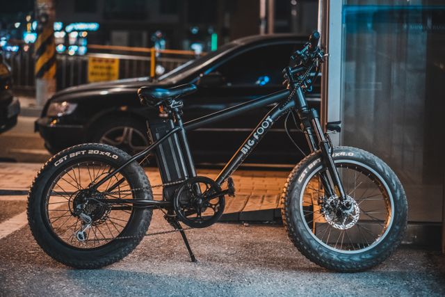Black Bigfoot electric fat-tire bicycle parked on a city street at night, streetlights blurred behind.